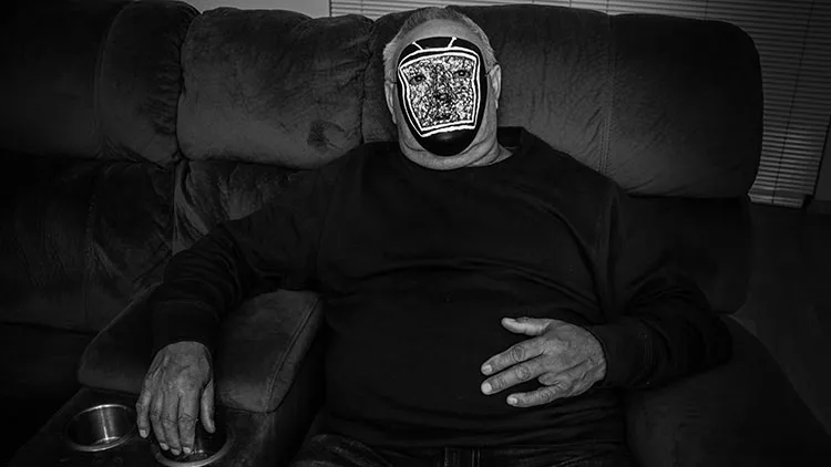 Man wearing a mask with a static TV painted on it. The man is sitting on the couch with the glow of a TV on them.