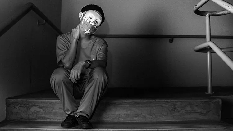 Man wearing a mask with heartbeat wave on it. He appears to be a surgeon sitting on some stairs in a dark stairway looking exhausted. A dark shadow is behind them.