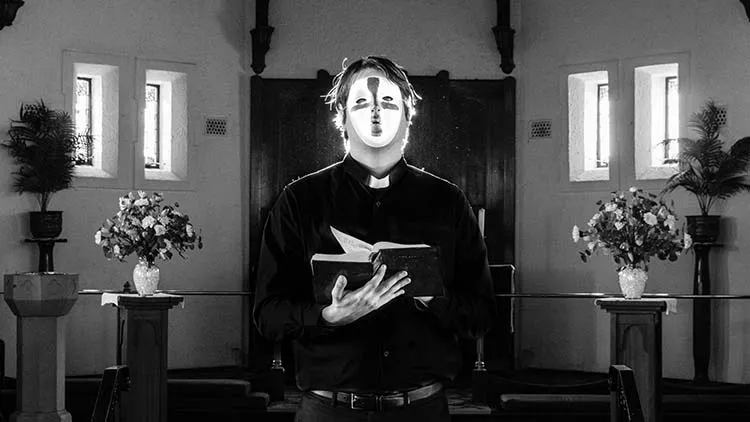 Priest with a mask with a cross on it standing in a church while reading the Bible.