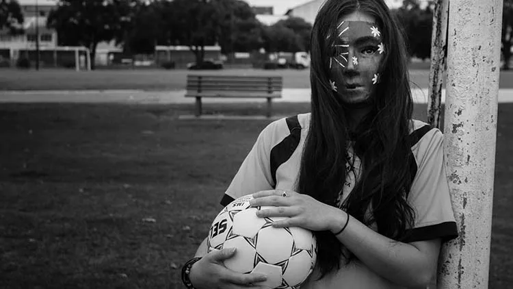 Woman with a mask of the Australian flag holding a soccer ball and standing next to a goal.