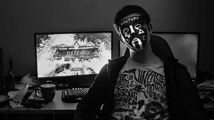 Man with a mask of various gaming symbols sitting in his room after clearly losing a videogame in the background.