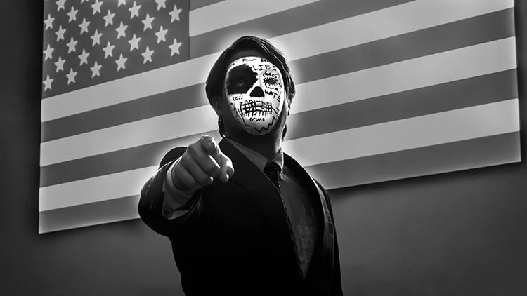 Man with a mask that is styled like a skull with various words like hate and lies standing infront of the American flag and pointing at the viewer.