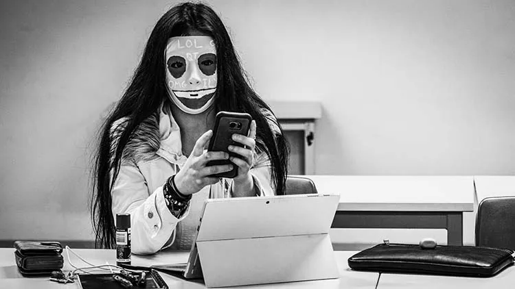 Female student wearing a mask with various meme culture words, sitting in a classroom and scrolling on their phone.