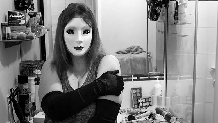 Woman standing in their bathroom wearing a mask with make-up and fake lashes on it. The woman looks nervous.