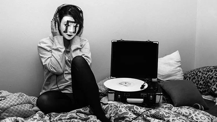 Girl wearing a mask with a musical symbol on it. The girl is in their Room wearing headphones plugged into a digital vinyl player.