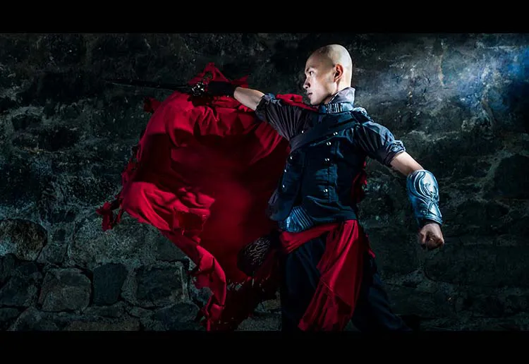 Man swinging a sword and is wearing an outfit inspired by high fantasy costumes. The man is wearing a red cape and a gauntlet with a money bag on his waist and a dark top and pants.