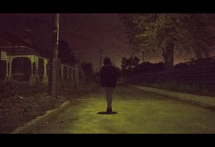 Eerie Image of a human figure walking down a side road near an abandoned building at night. The image has a yellow and red grainy hue.