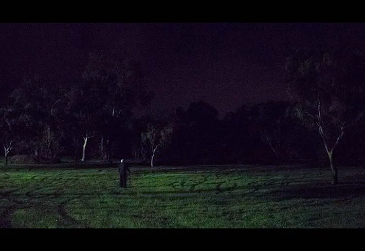 Image looking from afar at an old man using a cane who is staring up at the stary sky in a wide field.