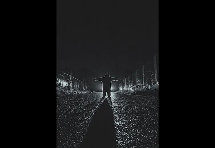 Black and white image of a figure kneeling with their arms spread wide. The light in front of them lights up the street the kneel in leaving a large shadow.