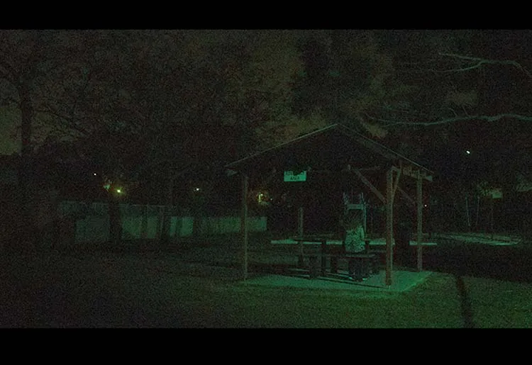 A green tinted image of a humanoid figure siting under a gazebo covering sitting straight up late at night.