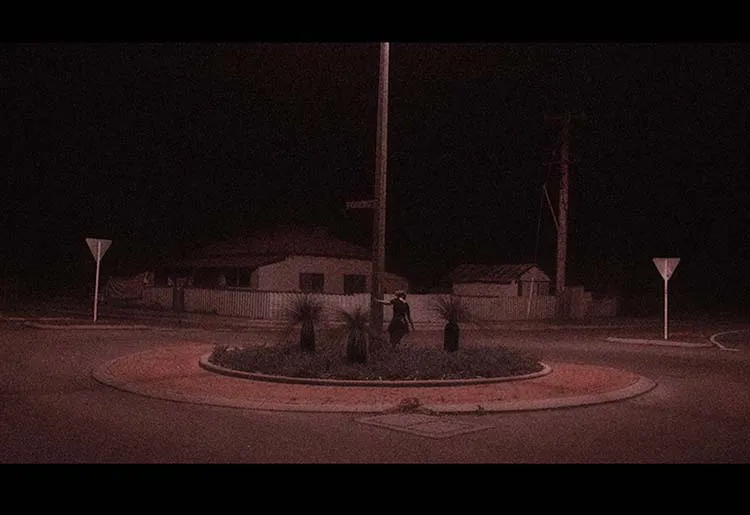 A red tinted image of a female figure swinging around a street pole which solely illuminates the scene.